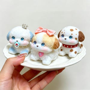 May include: Three small needle-felted puppy figurines with large blue eyes. The left puppy has a crown, pacifier, and pearl necklace. The center puppy wears a pink bow and a heart pendant. The right puppy has brown spots and a red studded collar.