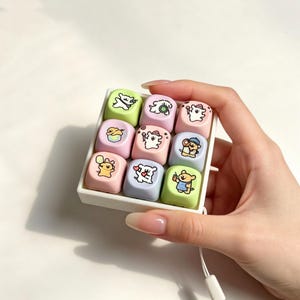 Pode incluir: Uma bandeja quadrada branca contendo nove teclas mecânicas individuais para teclado em tons pastel de rosa, azul e verde. Cada tecla apresenta uma pequena ilustração única de personagem de desenho animado, incluindo ursos e outros animais.
