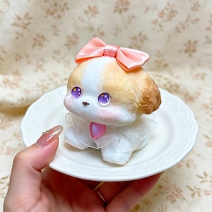 May include: A small, sculpted puppy figurine with white and tan fur, large purple eyes, and pink cheeks. The puppy wears a peach-colored satin bow on its head and a white sheer ruffled collar with a small pink heart-shaped charm at the center.