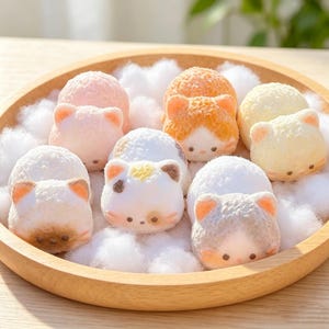 May include: A collection of small, round, cat-shaped confections in various pastel colours including pink, orange, yellow, and white, arranged on a circular wooden tray nestled among soft, white, fluffy material resembling clouds or cotton.