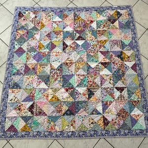 May include: A patchwork quilt with a geometric design. The quilt features numerous triangles in various floral patterns and solid colours, including pink, purple, and yellow. A floral patterned border in shades of blue and purple frames the quilt.