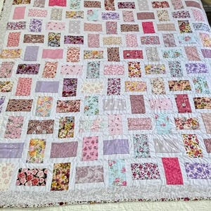 May include: A patchwork quilt featuring a white base and a multitude of rectangular patches. These patches showcase diverse floral and solid patterns in pinks, purples, and blues. The quilt has a patterned border, creating a vibrant and inviting aesthetic.