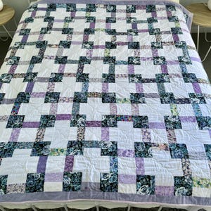 May include: A patchwork quilt featuring a white base with a grid design of floral and solid blocks in purple, blue, and green hues. The quilt is placed on a bed, complemented by white pillows and a pale purple border. The design is handmade.