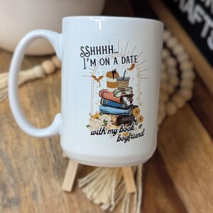 May include: White ceramic mug with a black and gold design. The mug features a stack of books, coffee cups, and the text "Sshhh... I'm on a date with my book boyfriend."