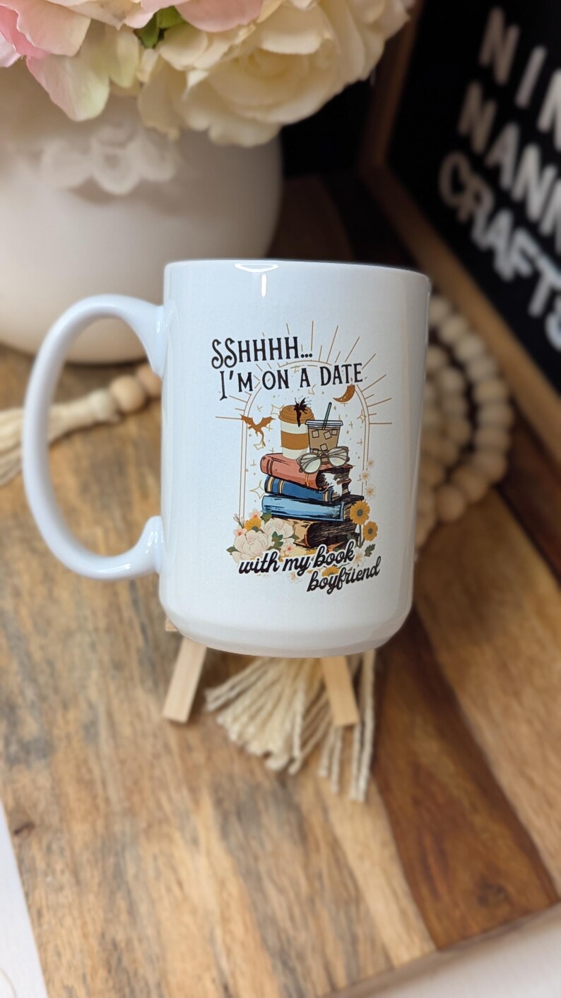 May include: White ceramic mug with a black and gold design. The mug features a stack of books, coffee cups, and the text "Sshhh... I'm on a date with my book boyfriend."
