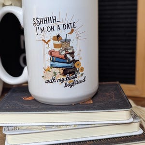 May include: White ceramic mug with a black and brown illustration of a stack of books, a coffee cup, and a pair of glasses. The text on the mug reads "Ssshhh... I'm on a date with my book boyfriend."