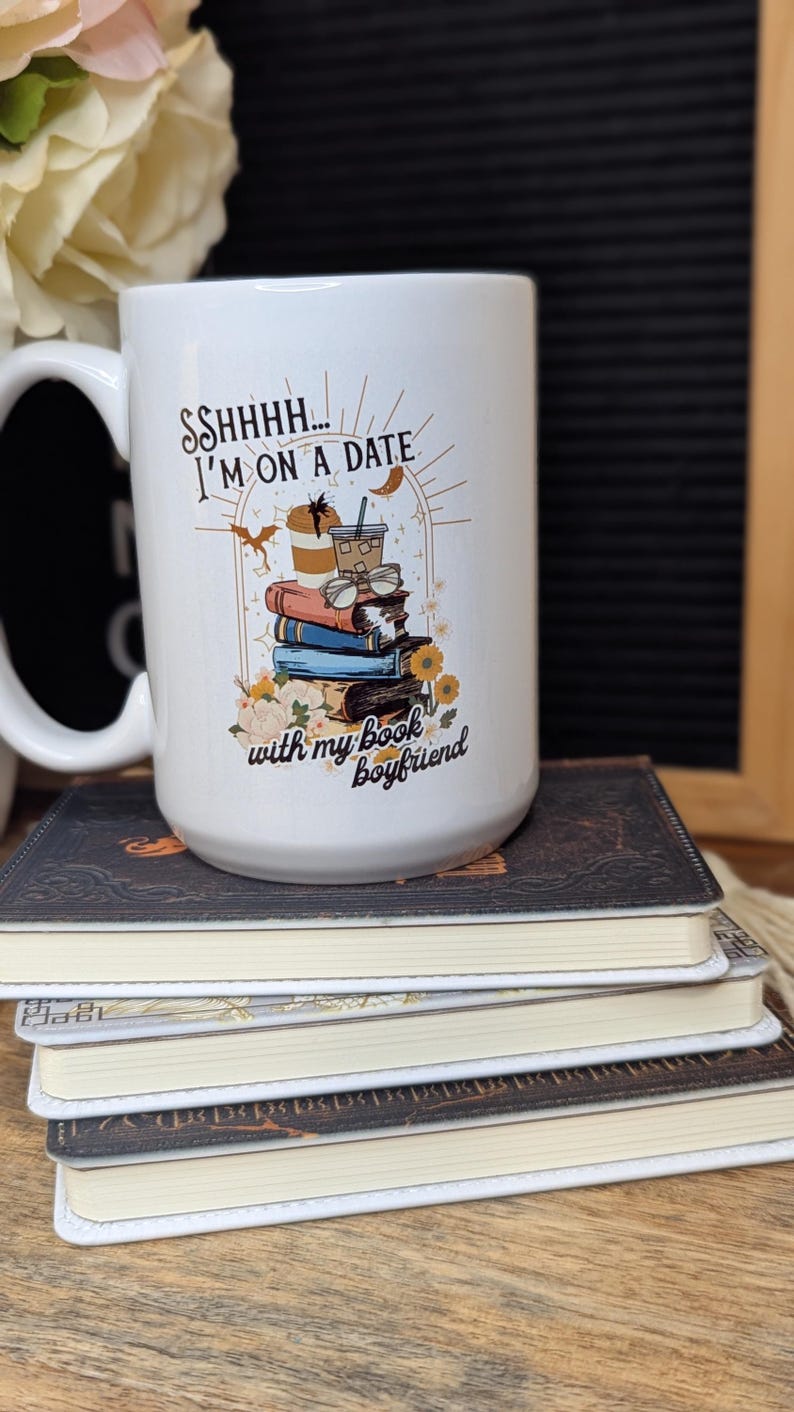May include: White ceramic mug with a black and brown illustration of a stack of books, a coffee cup, and a pair of glasses. The text on the mug reads "Ssshhh... I'm on a date with my book boyfriend."