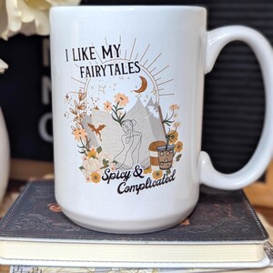 May include: White ceramic mug with a brown graphic that says "I like my fairytales spicy & complicated". The graphic features a woman sitting with a book, a dragon, flowers, and a cup of coffee.