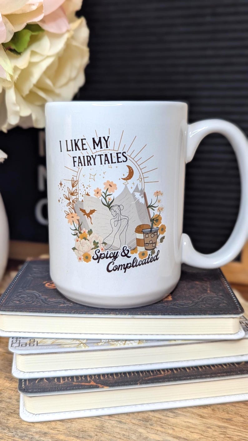 May include: White ceramic mug with a brown graphic that says "I like my fairytales spicy & complicated". The graphic features a woman sitting with a book, a dragon, flowers, and a cup of coffee.