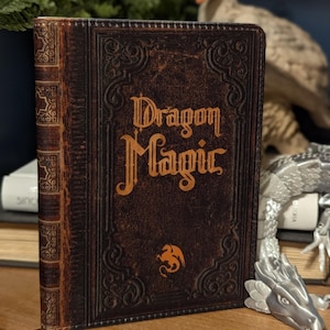 May include: A hardcover book titled "Dragon Magic" with a brown leather cover and gold lettering. The book has an embossed dragon emblem and a built-in bookmark. The book is displayed on a wooden surface with decorative items in the background.