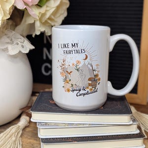 May include: White ceramic mug with a black and brown illustration of a mountain scene with the text "I like my fairytales spicy & complicated".