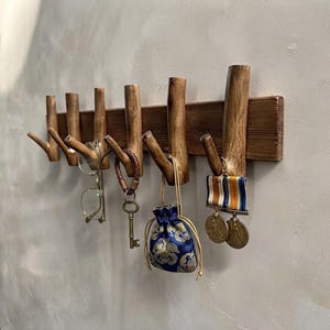 May include: A wall-mounted wooden coat rack featuring six natural branch-style hooks attached to a horizontal rectangular board. The hooks hold various items including a pair of glasses, a metal key on a leather strap, a small blue pouch, and medals.