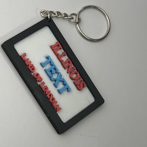 Op de afbeelding: Een rechthoekige sleutelhanger met een zwart frame en een wit midden. De sleutelhanger is voorzien van verhoogde rode letters met de tekst ILLINOIS en LAND OF LINCOLN, en verhoogde blauwe letters met de tekst TEXT, bevestigd aan een zilveren metalen ring.