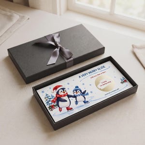 Puede incluir: Una caja de regalo rectangular negra con una cinta gris, que contiene una tarjeta blanca con el texto A VERY MERRY GLIDE, With love, Mum & Dad, y Scratch for A Christmas gift. La tarjeta muestra dos pingüinos ilustrados patinando.