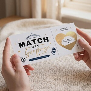 May include: A white rectangular ticket with black and gold text reading MATCH DAY Surprise and YOU'RE OFF TO THE GAME. It features a soccer ball, a stadium arch, a scarf, and a gold heart-shaped scratch-off area labeled SCRATCH TO REVEAL.