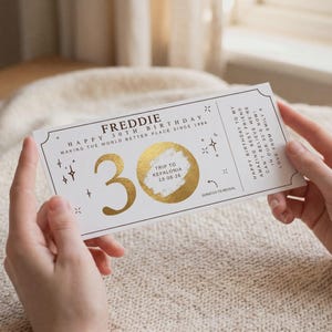 May include: A white birthday scratch card with gold foil detailing. The card is for a 30th birthday and includes the name "Freddie". It features a scratch-off area for a trip to Kefalonia. The card is held in two hands.