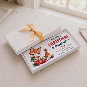 Puede incluir: Una caja de regalo rectangular blanca con un lazo dorado. Dentro, una tarjeta muestra un zorro en un calcetín navideño rojo, un petirrojo con un regalo y el texto A LITTLE CHRISTMAS MISCHIEF, Isla, For our favourite little fox, Christmas 2026, With love, Mum & Dad xxx.