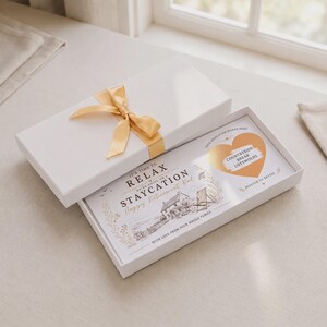 Könnte beinhalten: Eine weiße rechteckige Geschenkbox mit goldener Schleife enthält eine Ruhestands-Geschenkkarte. Die Karte zeigt eine Cottage-Illustration, den Text 'IT'S TIME TO RELAX' und ein goldenes herzförmiges Rubbelfeld mit 'COUNTRYSIDE BREAK COTSWOLDS'.