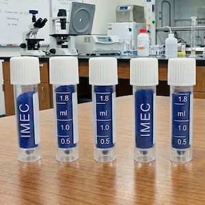 May include: Five clear plastic vials with white caps, each containing blue powder. The vials have a blue label with the text "IMEC" and volume markings of 0.5, 1.0, and 1.8 ml. The vials are arranged on a wooden surface.