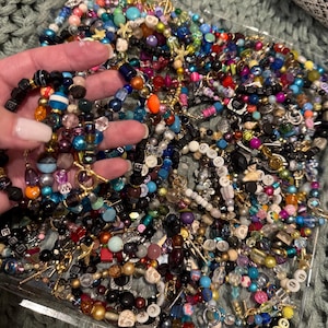May include: A collection of beaded bracelets in various colors and designs. The bracelets feature a mix of beads, including pearls, glass beads, and letter beads. Some bracelets have gold-colored clasps and charms. The bracelets are displayed on a gray surface.
