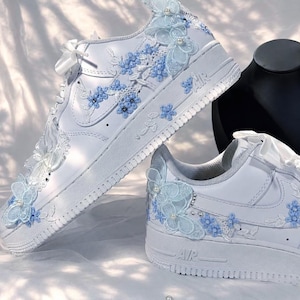 Puede incluir: Un par de zapatillas blancas con aplicaciones de encaje floral azul claro, adornos de mariposas transparentes y pequeñas perlas blancas. Los zapatos tienen cordones de cinta de satén blanco y el texto AIR grabado en las suelas gruesas.