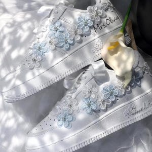 Puede incluir: Un par de zapatillas blancas con cordones de cinta de satén y aplicaciones de encaje floral en tonos blanco y azul claro. Cada flor está acentuada con una cuenta de perla central. El texto Mrs. Olivia está escrito en el lateral de un zapato.