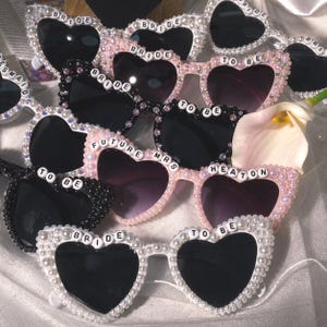 Puede incluir: Gafas de sol en forma de corazón en blanco, rosa y negro, adornadas con perlas y pedrería. Algunas gafas presentan texto como "BRIDE", "TO BE" y "FUTURE MRS HEATON" en las monturas. Las lentes son oscuras.