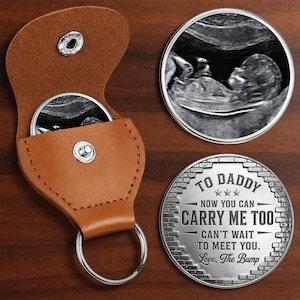 Puede incluir: Un llavero de cuero marrón con un llavero plateado, que contiene una moneda redonda con una imagen de ultrasonido. Una moneda de plata separada dice "TO DADDY, NOW YOU CAN CARRY ME TOO, CAN'T WAIT TO MEET YOU. Love, The Bump."
