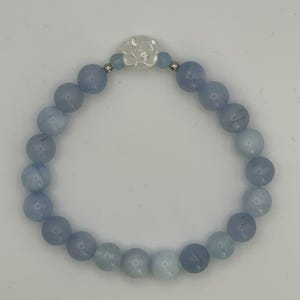 May include: A beaded bracelet featuring round, light blue, semi-translucent stone beads arranged in a circular pattern. At the top center, a single white, carved, flower-shaped bead is flanked by two small, metallic silver-toned spacer beads.