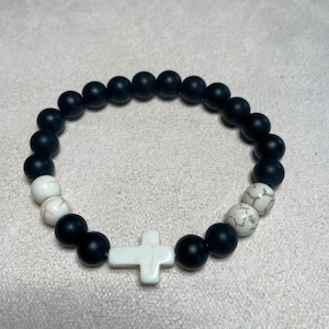 May include: A stretch bracelet featuring matte black round beads, two white marble-patterned round beads on one side, and a white marble-patterned cross-shaped bead centred at the bottom, with a continuous circular design.