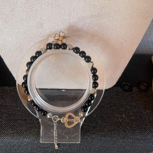 May include: A beaded bracelet featuring round black beads interspersed with small silver-toned metal charms. The charms include heart shapes and a four-leaf clover. The bracelet has a silver-toned metal toggle clasp shaped like a heart.