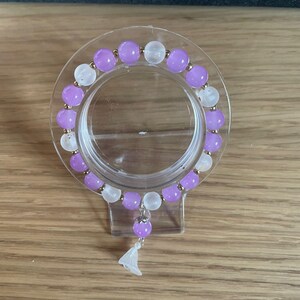 May include: A beaded bracelet featuring alternating round purple and translucent white beads separated by small gold-toned spacers. A single purple bead hangs from the bottom, attached to a small white bell-shaped charm with a silver-toned metal bail.