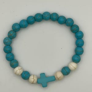 May include: A circular stretch bracelet composed of round turquoise-colored beads with dark veining, interspersed with a few smooth white beads and a single turquoise-colored cross-shaped bead positioned at the bottom of the circular arrangement.