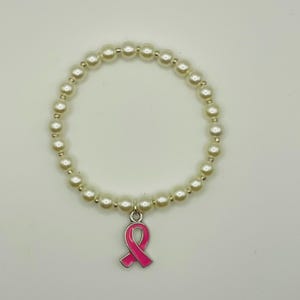 May include: A circular stretch bracelet composed of smooth, cream-colored faux pearl beads. The bracelet features a single metal charm in the shape of a awareness ribbon, finished with a glossy, vibrant pink enamel coating on the front surface.