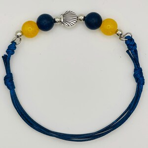 May include: A bracelet featuring a blue cord with knots, two yellow beads, two dark blue beads, and a central silver-toned metal shell charm. The design includes small silver-toned spacer beads between the larger colored beads on the cord.