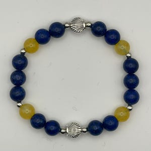 May include: A beaded stretch bracelet featuring alternating dark blue and translucent yellow round beads. The design includes two silver-toned metal seashell charms positioned opposite each other, accented by small silver-toned spacer beads.