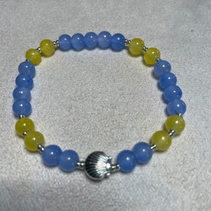 May include: A beaded bracelet featuring alternating light blue and yellow round beads separated by small silver-toned spacers. The bracelet includes a central silver-toned metal charm shaped like a seashell, creating a circular design.