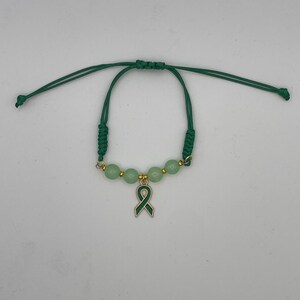 May include: A green cord bracelet featuring four light green round beads separated by small gold-toned metal spacers. A central dark green awareness ribbon charm hangs from the middle, and the bracelet has adjustable knotted cord ends.