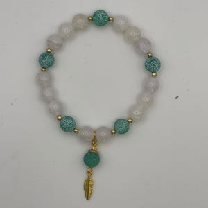 May include: A beaded bracelet featuring alternating pale pink translucent beads and teal crackle-textured beads. Small gold-toned metal spacer beads are placed between the larger beads. A single teal crackle bead and a gold-toned feather charm hang.