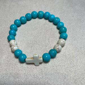 May include: A stretch bracelet featuring round turquoise-coloured beads with dark veining, interspersed with two white beads with grey veining on each side and a central white cross-shaped bead. The beads are arranged in a circular pattern.