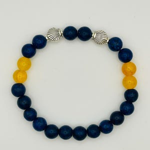 May include: A beaded bracelet featuring a circular arrangement of dark blue and yellow spherical beads. Two silver-toned metal charms shaped like seashells are positioned at the top of the bracelet, separating the blue beads from the yellow ones.