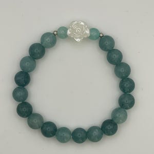 May include: A stretch bracelet composed of round, translucent teal-green gemstone beads. The bracelet features a central white, carved flower-shaped bead flanked by two smaller, lighter green beads and two small silver-toned metal spacer beads.