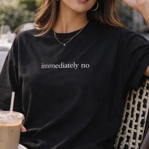 Puede incluir: Una taza de cerámica blanca con un asa negra y el texto "But First, Coffee" en negrita negra. La taza se encuentra sobre un fondo neutro, resaltando su diseño simple.