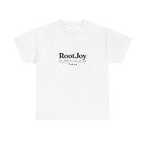 Può includere: Una t-shirt bianca a maniche corte con il testo nero RootJoy centrato sul petto. Sotto il testo si trova una piccola grafica di un ramo di vite orizzontale, con la parola Tokyo stampata in un carattere più piccolo sotto il ramo.