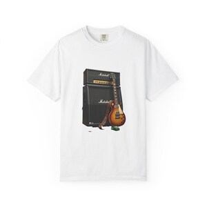 May include: A white short-sleeve t-shirt featuring a graphic print of a sunburst electric guitar leaning against a black Marshall amplifier stack. A brown leather guitar strap hangs from the guitar neck, and a small green effects pedal sits below.