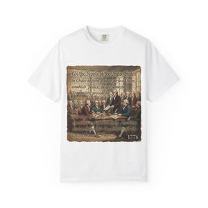Puede incluir: Camiseta blanca con una imagen de la firma de la Constitución de los Estados Unidos. La imagen incluye texto de la Constitución y el año 1776. La camiseta tiene cuello redondo.