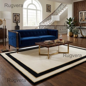 May include: A luxurious living room featuring a navy blue velvet sofa, a dark wood coffee table with gold legs, and a cream-colored rug with black and gold borders. The room has a grand staircase and elegant decor.