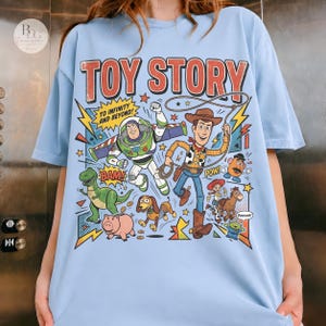 May include: Light blue t-shirt featuring a colorful graphic of Toy Story characters. The design includes Buzz Lightyear, Woody, and other characters with text that reads "TO INFINITY AND BEYOND!" and "TOY STORY".