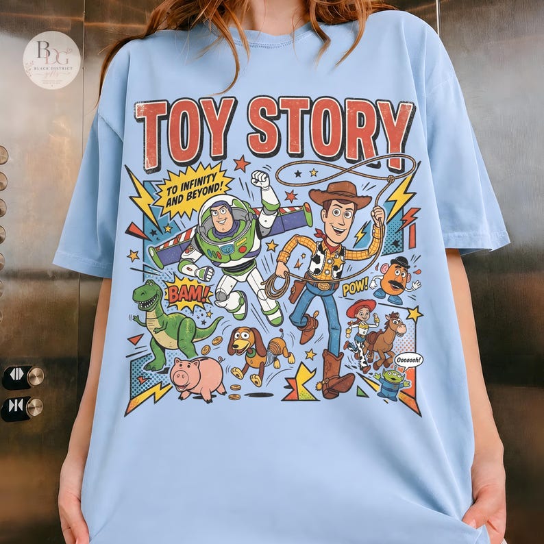 May include: Light blue t-shirt featuring a colorful graphic of Toy Story characters. The design includes Buzz Lightyear, Woody, and other characters with text that reads "TO INFINITY AND BEYOND!" and "TOY STORY".