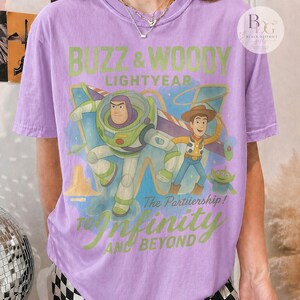 May include: A light purple t-shirt featuring Buzz Lightyear and Woody from Toy Story. The graphic includes the text "Buzz & Woody Lightyear" and "To Infinity and Beyond". The shirt has short sleeves.